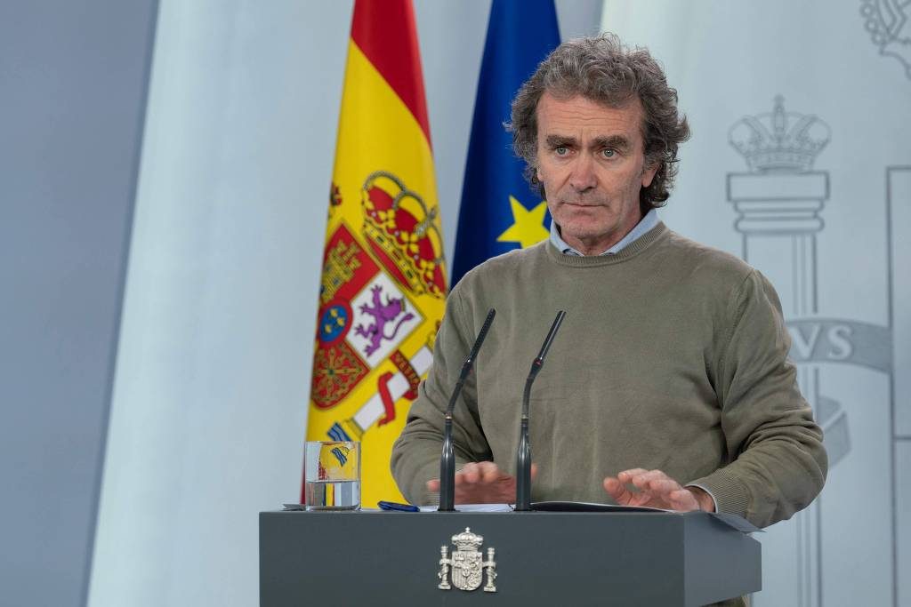 Fernando Simón fala à imprensa diante de um microfone, com a bandeira da Espanha e da União Europeia detrás